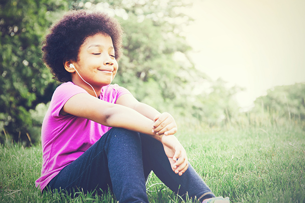 A young kid sits outside in the grass on a sunny day. Their eyes are closed and they are smiling while listening to someone using ear buds.