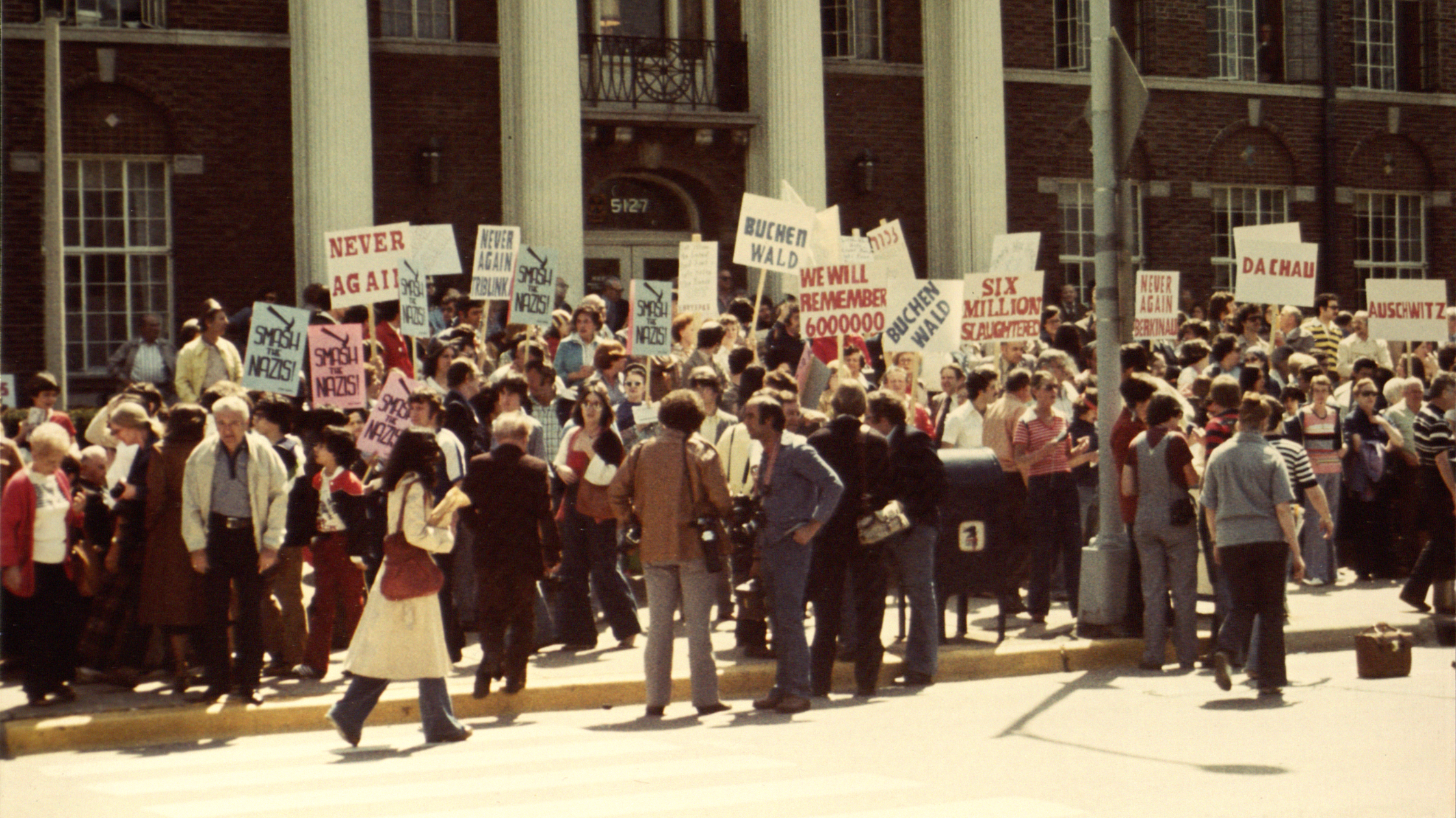 color photo of people protesting and holding signs in front of Village Hall. 