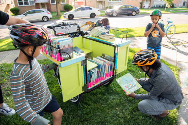 A woman crouches in front of the open cargo hold of the Skokie Public Library book bike. She holds up a book while her two children, two young boys, look on from either side of her. The three of them are wearing bike helmets. The cargo hold is yellowish-green and has dozens of books inside, most of which have a pink label on the spine.