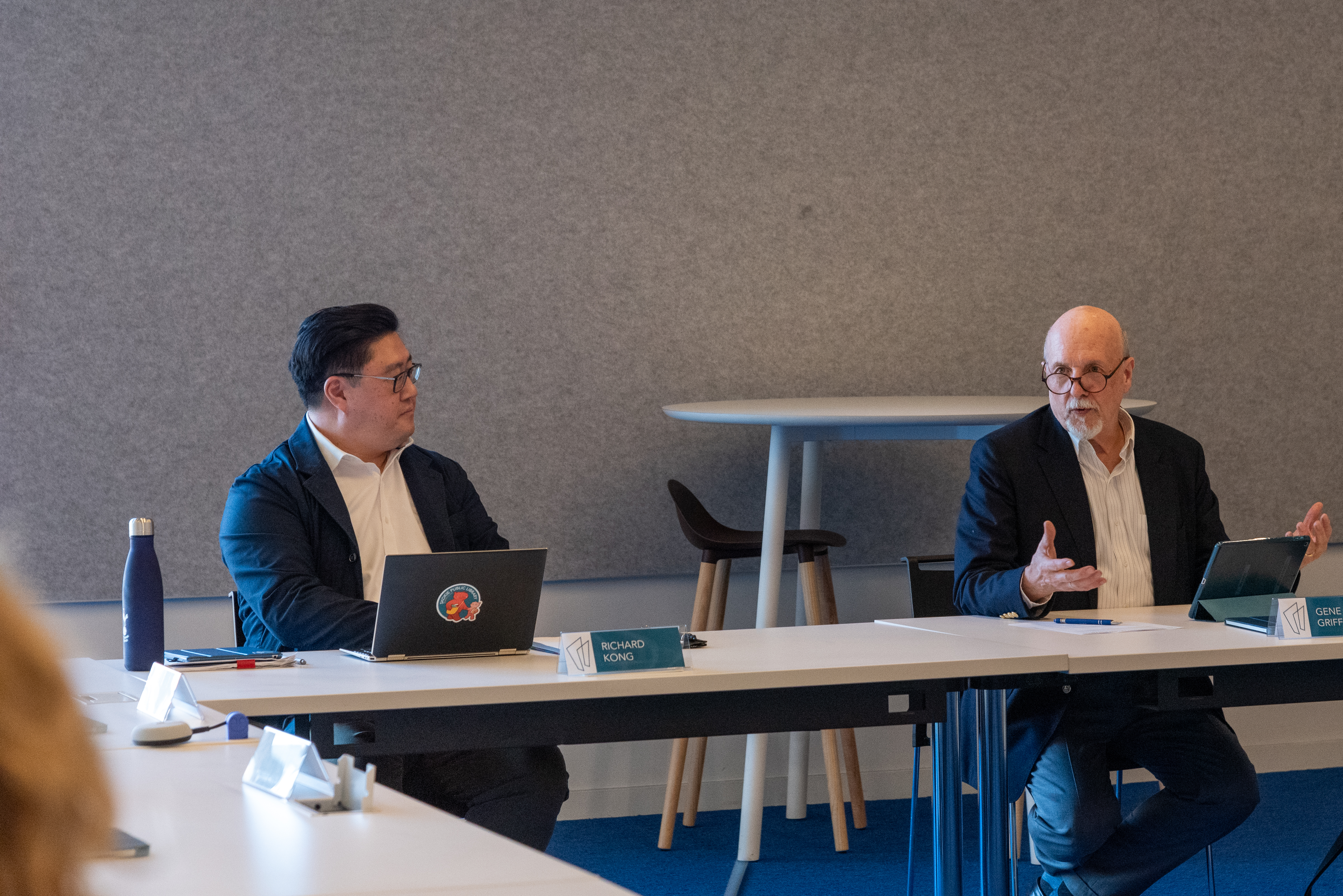 Two men seated at a table at the head of the room. The man on the right is speaking and gesturing.