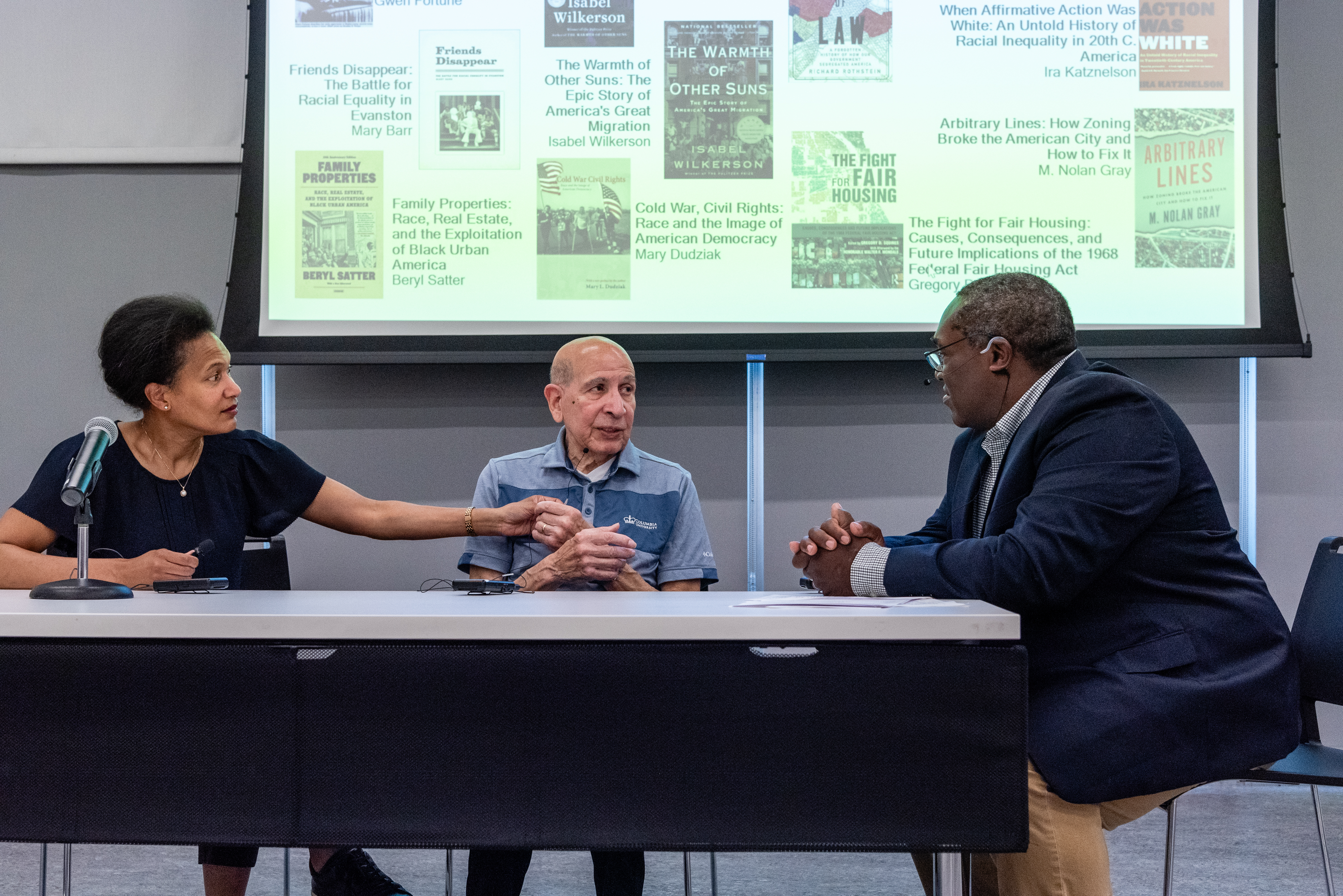 Two men and a woman sit at a table at the front of the room. The woman is grasping the hand of an older man. There is a powerpoint presentation behind them