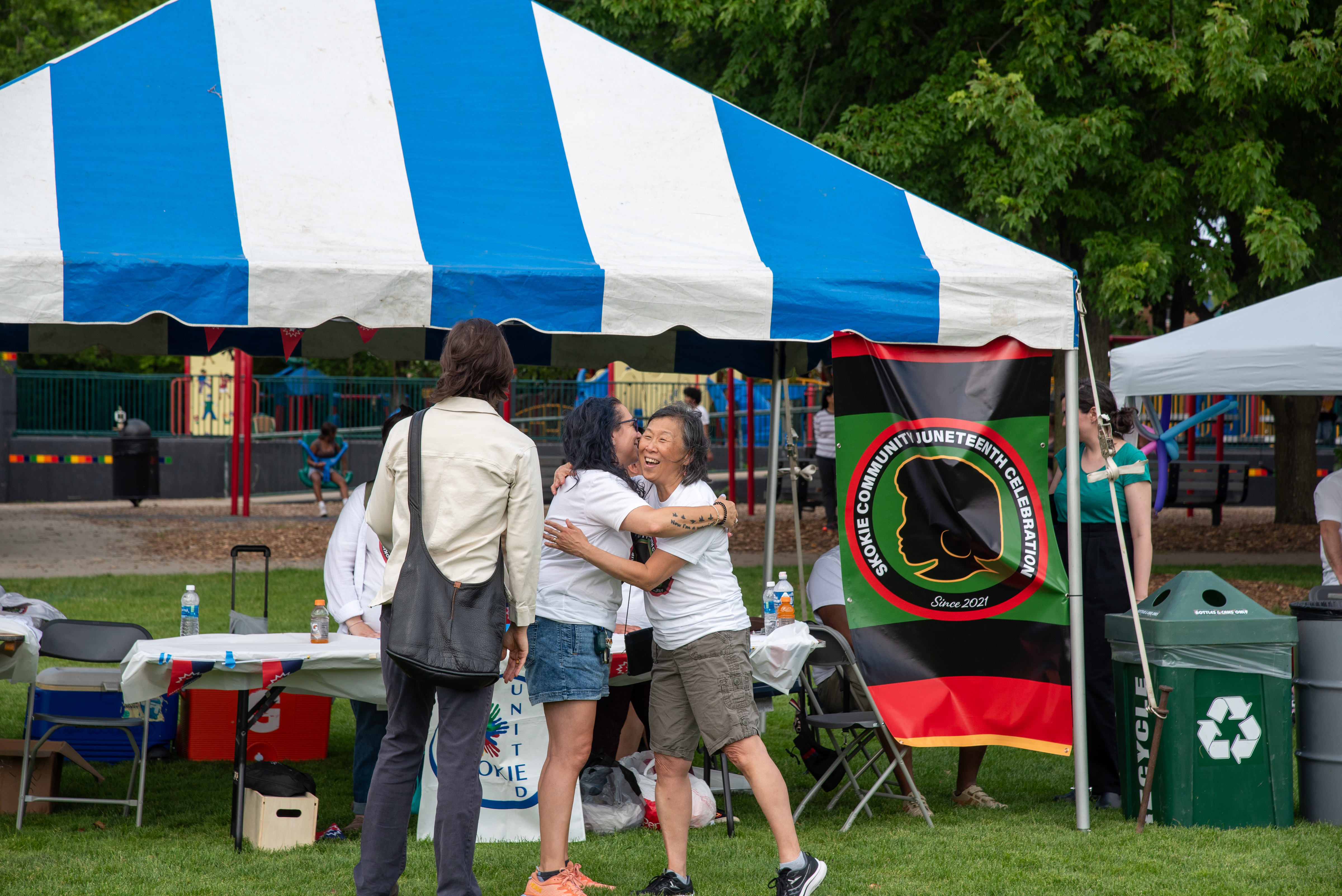 two people are embracing outside of an outdoor event tent. There is a flag of a Black person's silhouette in the colors black and red hanging from the tent