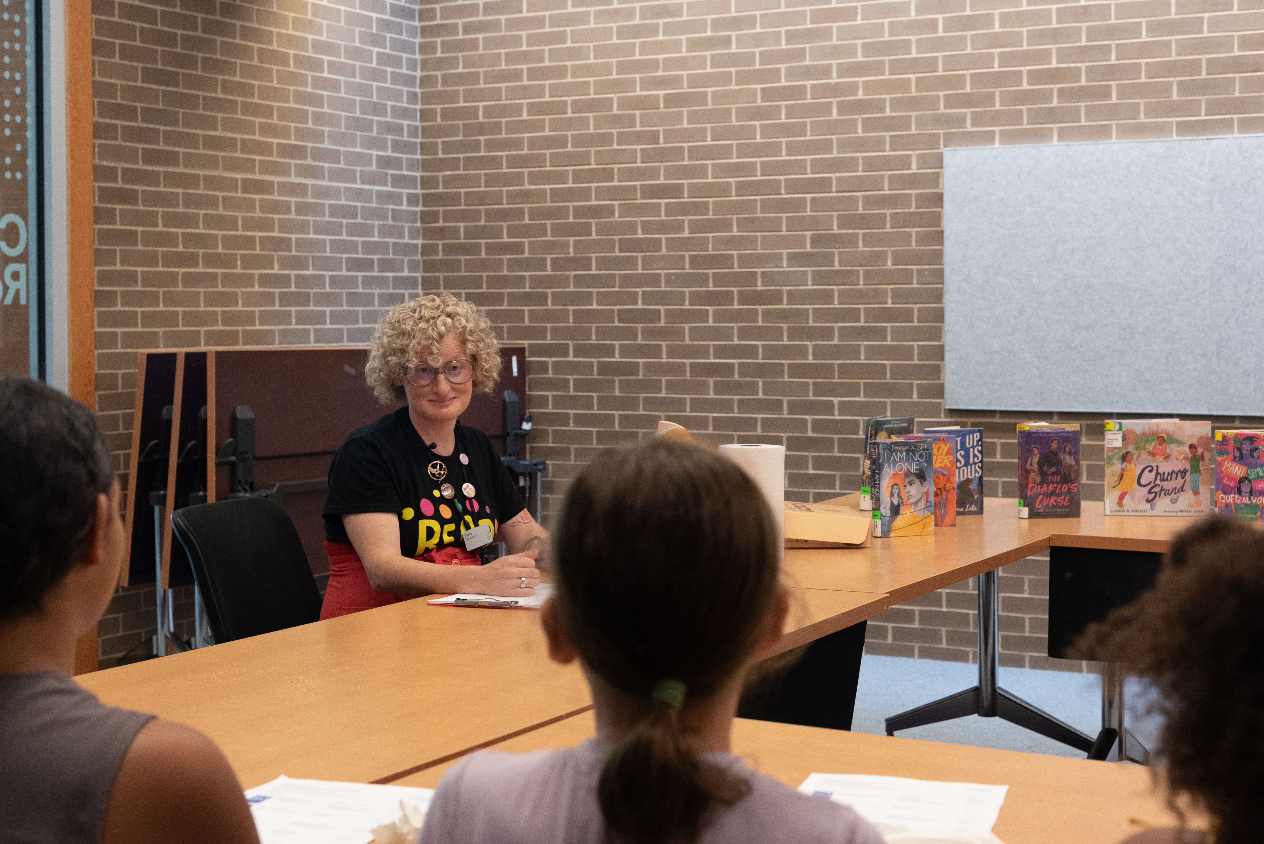 librarian sitting next to many young adult titles on a table while smiling at teens