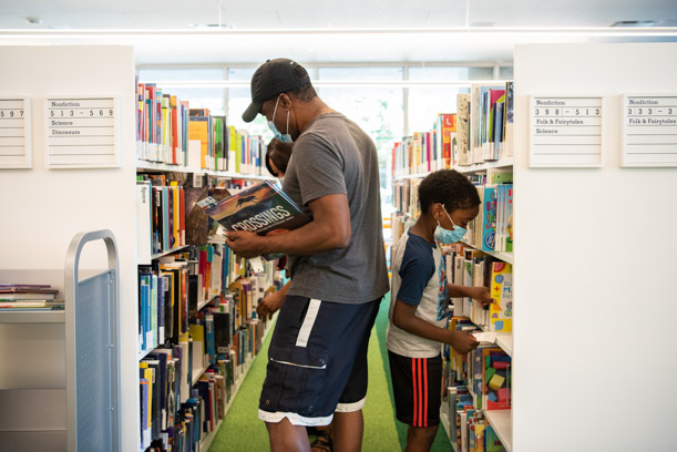 Three patrons, two adults and a child, stand back to back, browsing the shelves in the Kids Room.