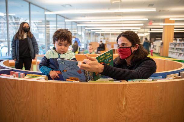 A woman leans on a circular shelf reading a board book to her child. The woman is wearing a red mask and the boy is wearing a blue jacket.