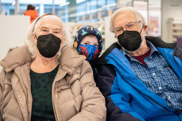 An older man and woman, both masked, pose for the camera with their grandson (also masked) who stands between them. The woman is wearing a green shirt and brown jacket, the man is wearing a blue shirt and blue jacket, and the child is wearing a hooded sweatshirt. 