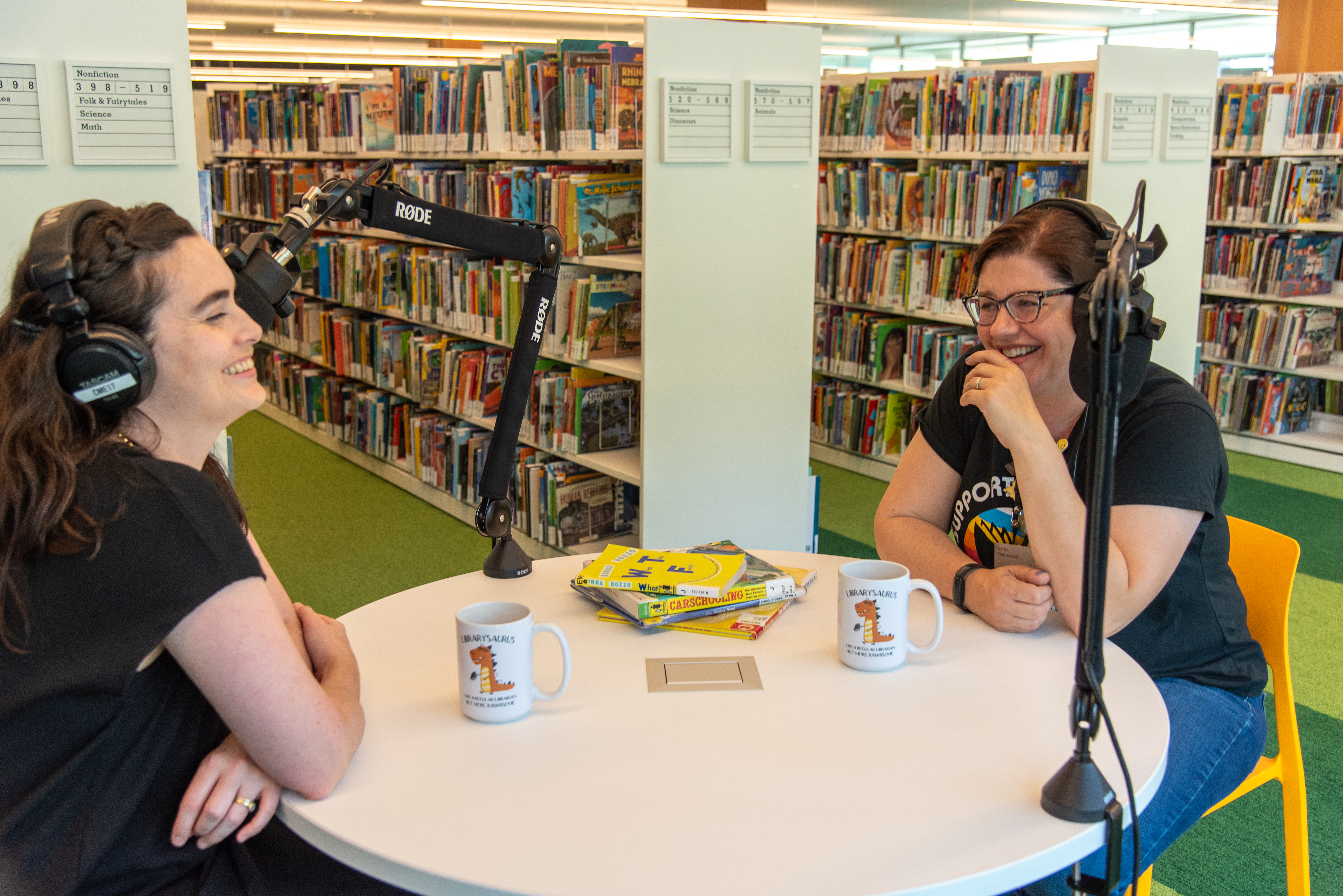 Katie and Caitlin are sitting a table in the Kids Room, laughing. They are facing each other, wearing headphones, and speaking into microphones. 