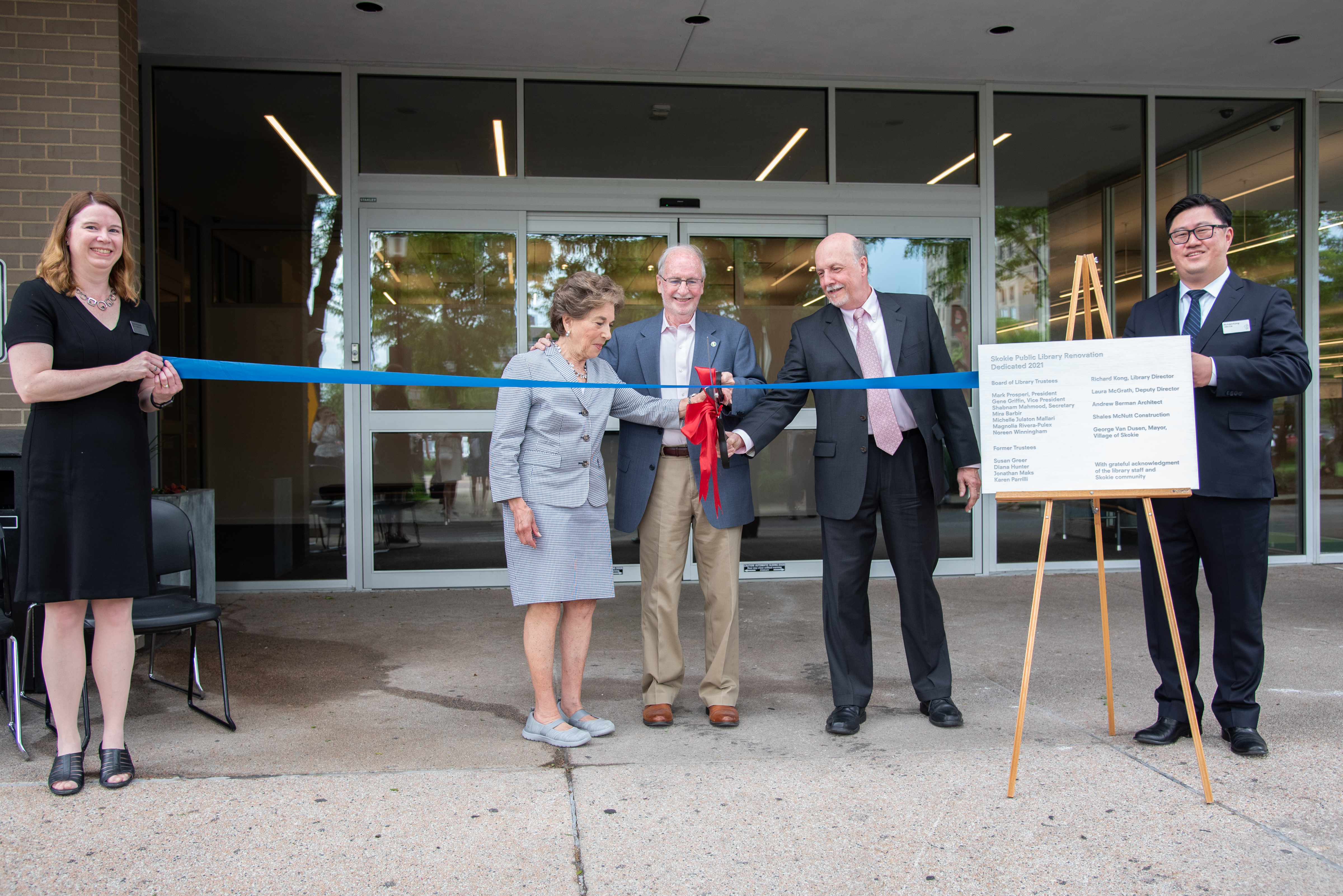 A group of three people wearing business suits use an oversized pair of scissors to cut a ribbon being held by two others