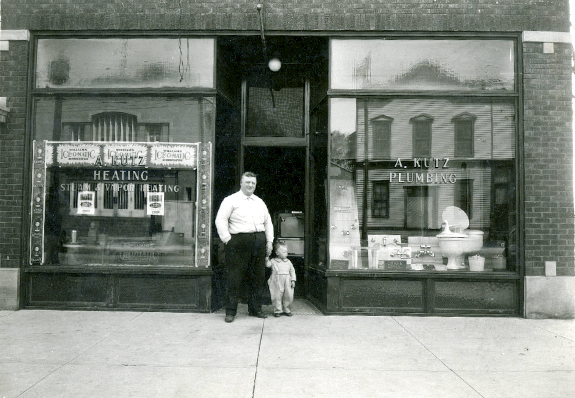 Black-and-white photograph of the A. Kutz Plumbing and Heating storefront in Niles Center, Illinois in 1910. An adult man and a toddler are standing in front of the building. Black-and-white photograph of the A. Kutz Plumbing and Heating storefront in Niles Center, Illinois in 1910. An adult man and a toddler are standing in front of the building.